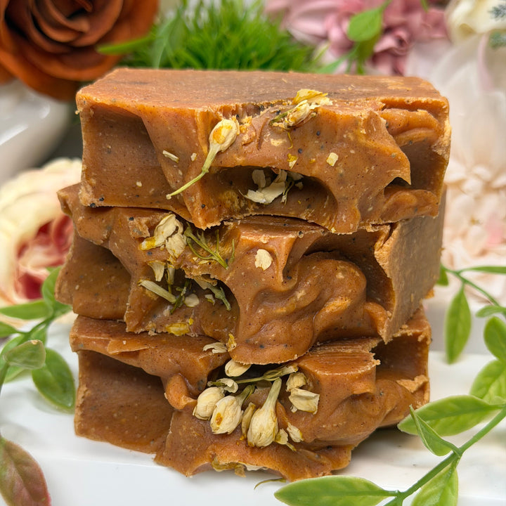 Brown soap bar with visible herbs on a white background with flowers and leaves.