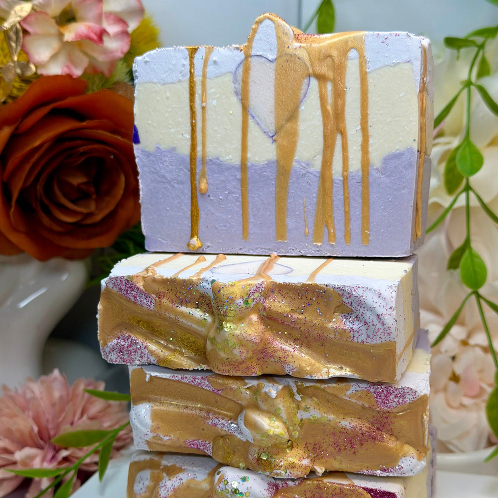 Stack of decorative soap bars with gold and purple drizzle on a white surface with flowers in the background