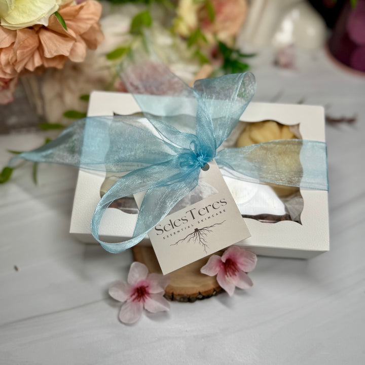 Gift box with a blue ribbon and a card, surrounded by flowers on a light surface.