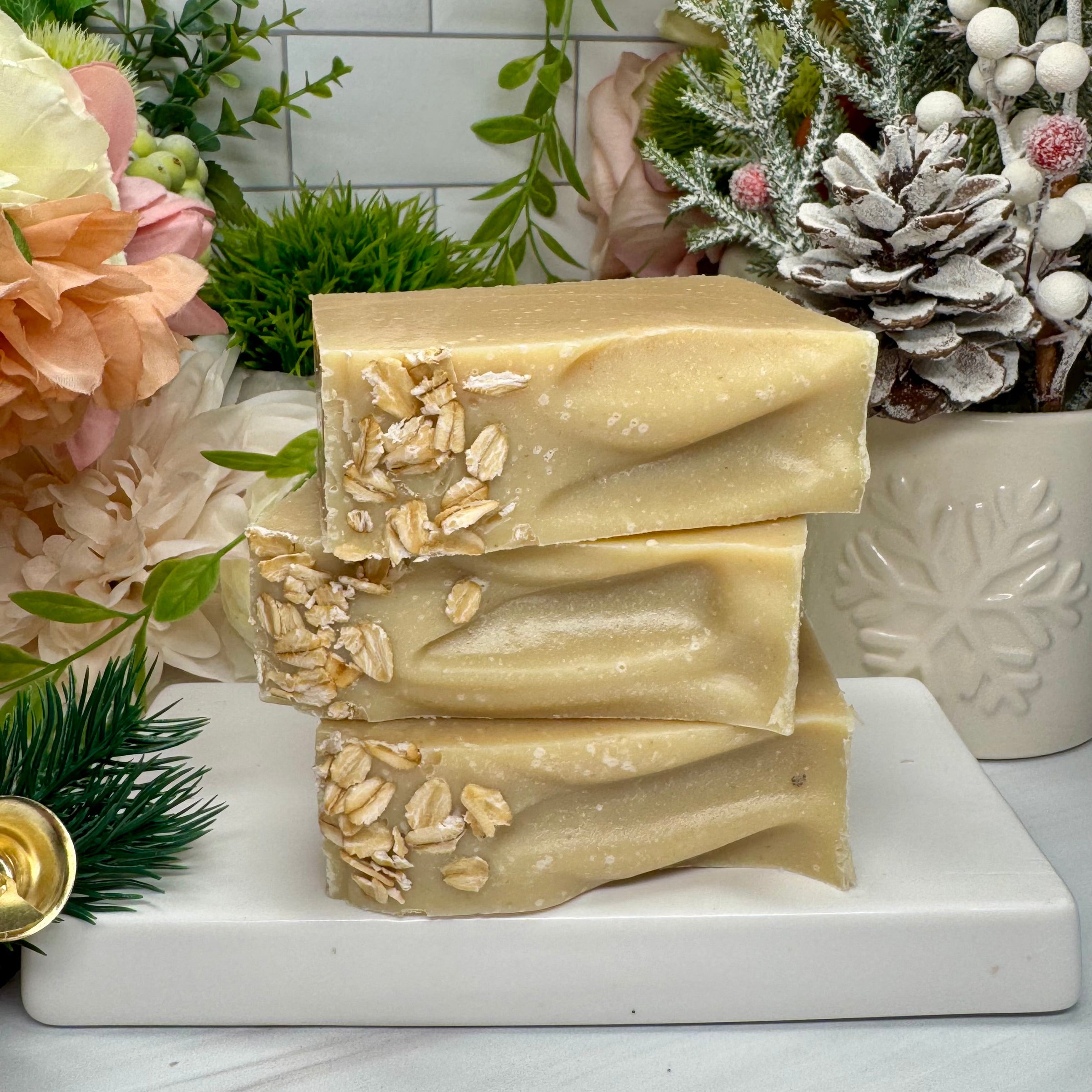Stack of natural soap bars with visible ingredients on a marble slab, surrounded by decorative plants and flowers.