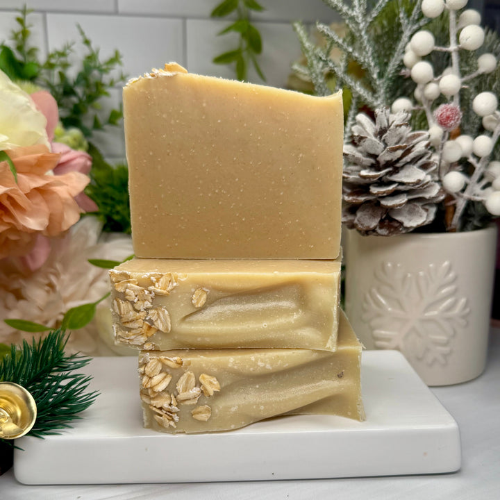 Three stacked soap bars with oatmeal on a marble stand with decorative plants in the background.