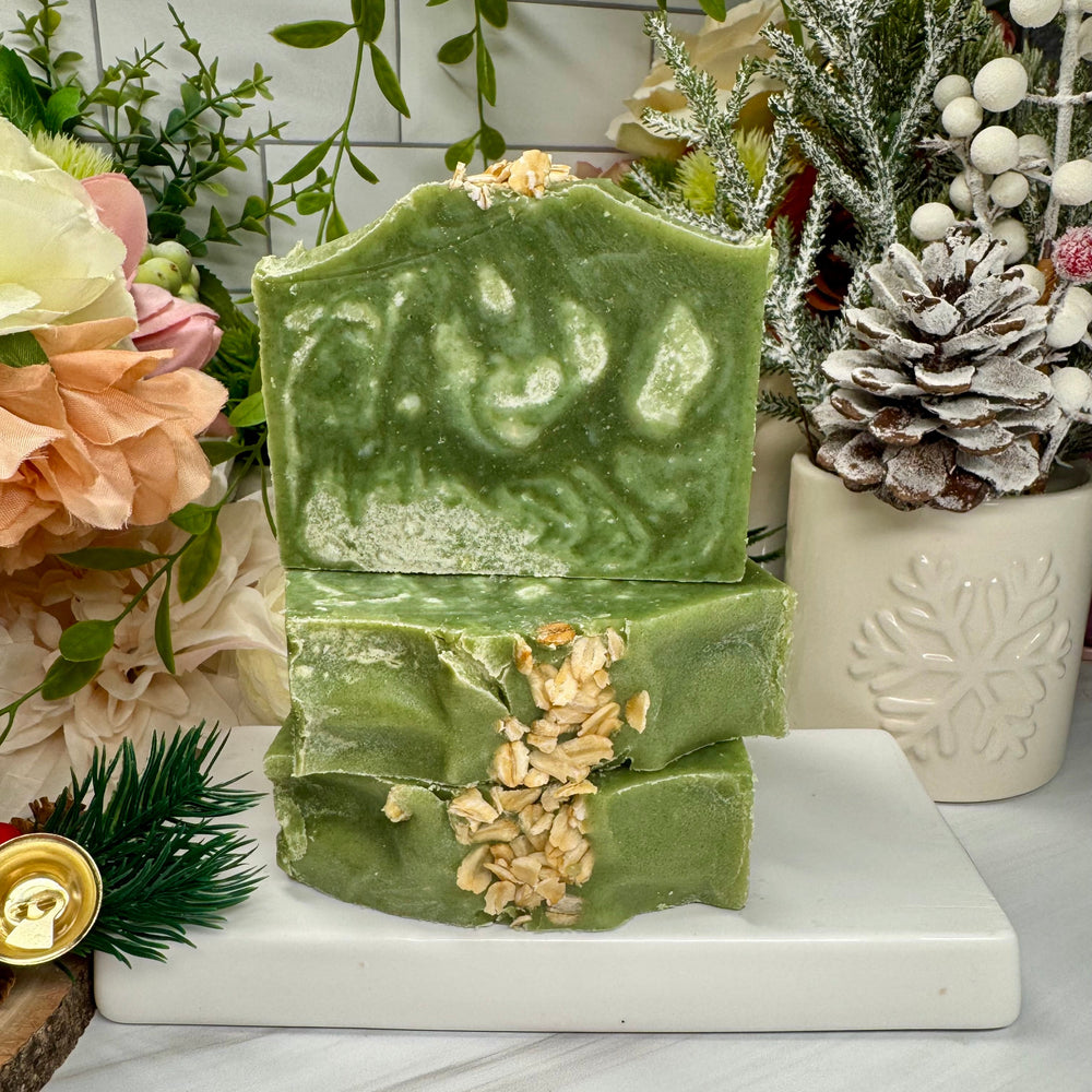 Green marbled soap bars with visible nuts on a white marble slab, surrounded by decorative plants and flowers.