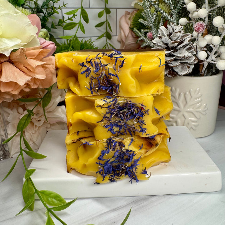 Stack of yellow soap with lavender petals on a white surface with decorative plants in the background.