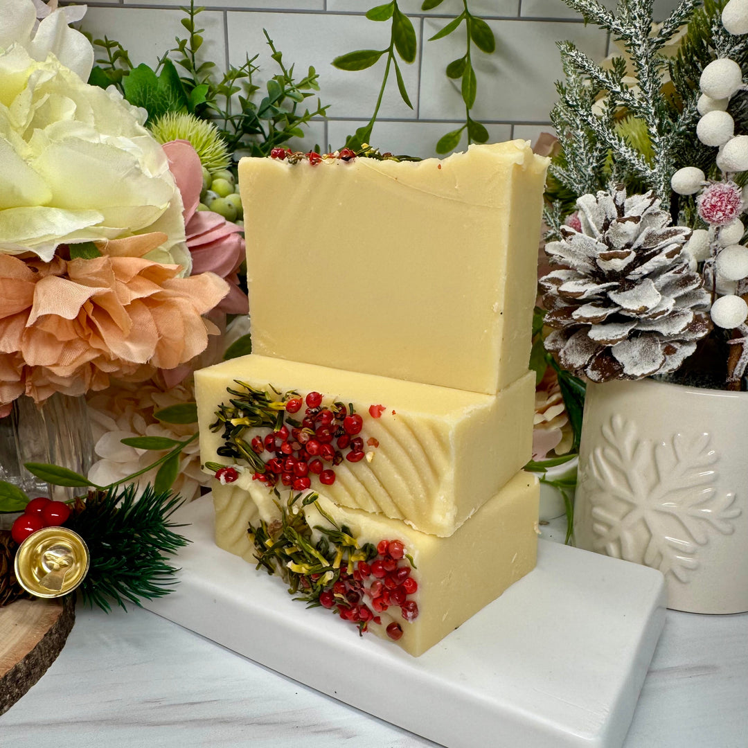 Two stacked blocks of cheese with decorative berries on a white surface, surrounded by flowers and a snowy pine cone.