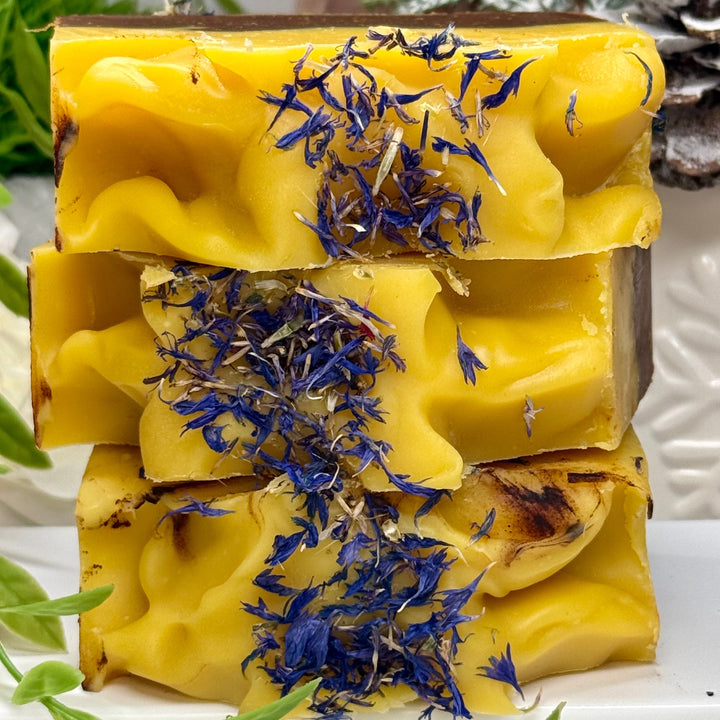 Three stacked yellow soap bars with blue dried flowers on a white surface with green leaves.