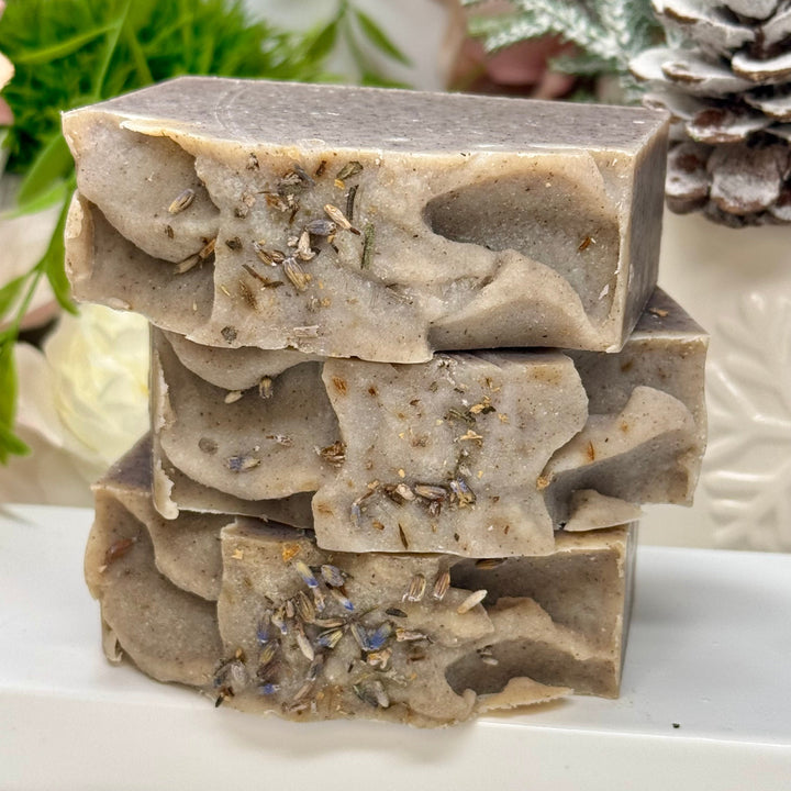 Stack of natural soap bars with visible herbs on a white surface, with plants and a decorative item in the background.