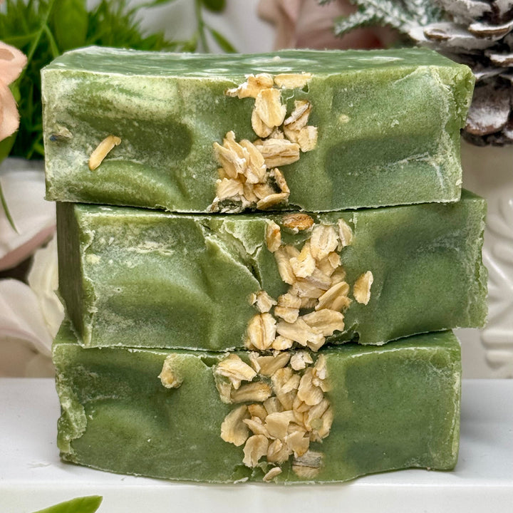 Stack of green soap bars with oatmeal pieces on a white surface with greenery in the background