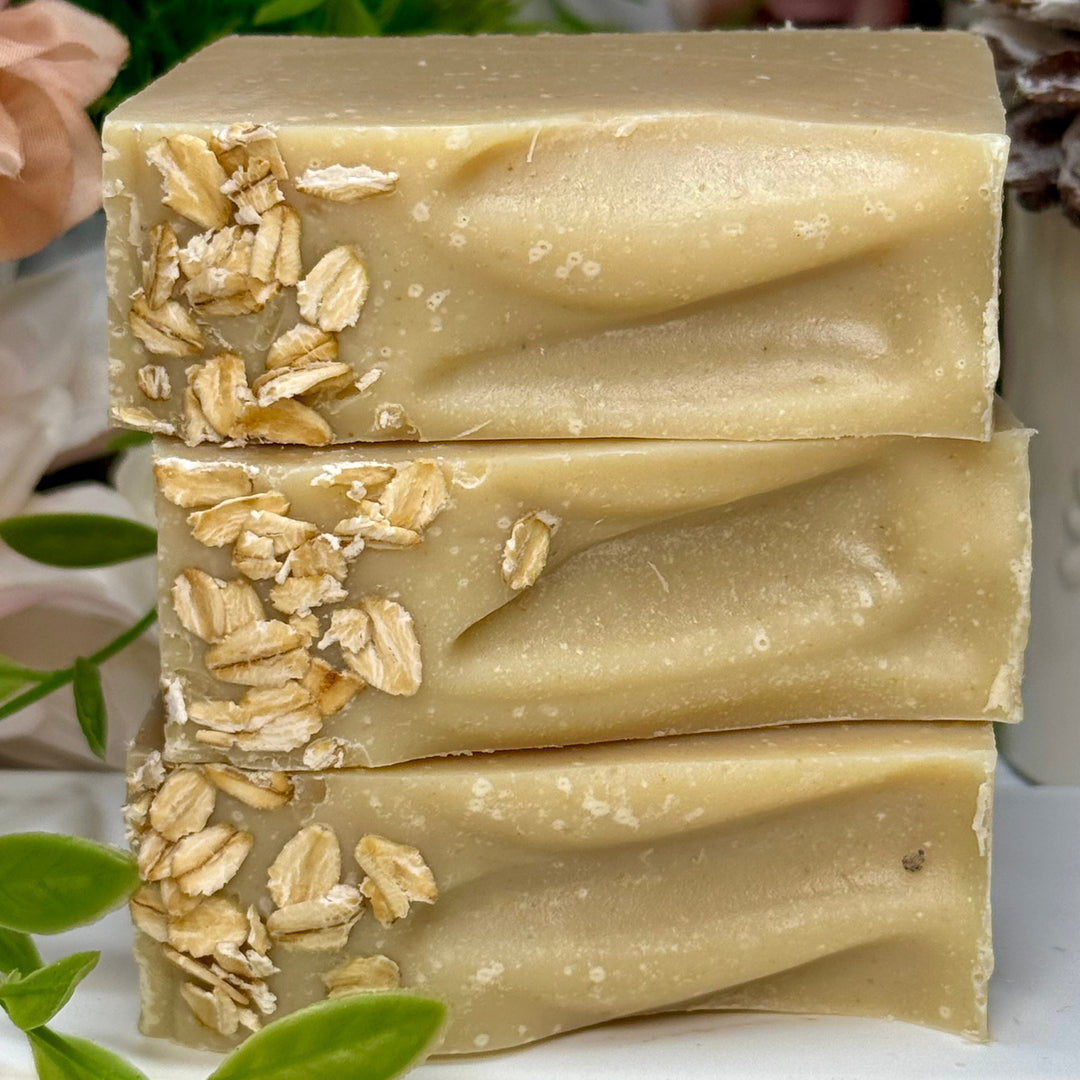 Three bars of oatmeal soap stacked on a white surface with greenery in the background